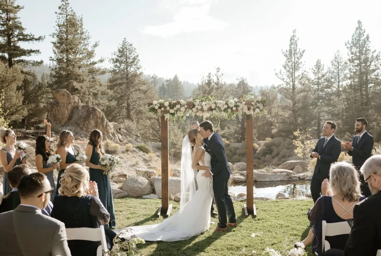 Couple kissing beneath the floral arch as the wedding party and seated guests look on
